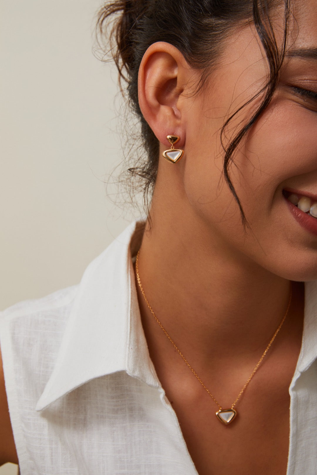 Woman wearing gold heart-shaped earrings and a necklace against a neutral background