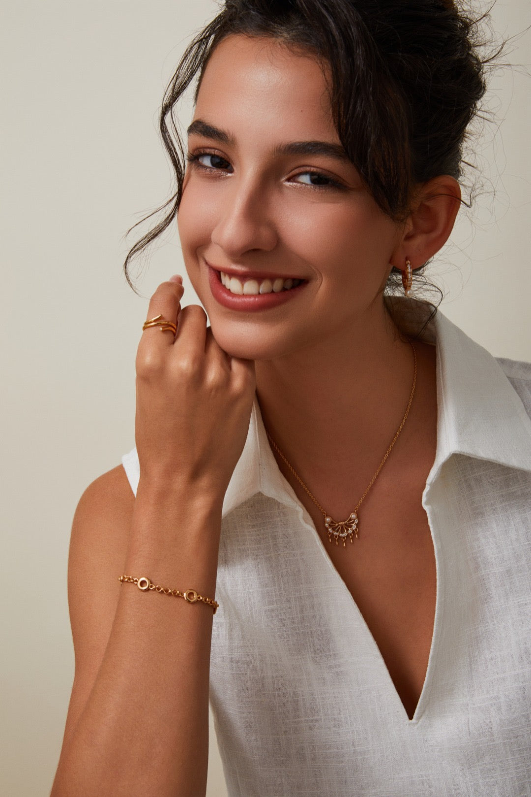 Woman wearing gold jewelry including a necklace, bracelet, and ring against a neutral background