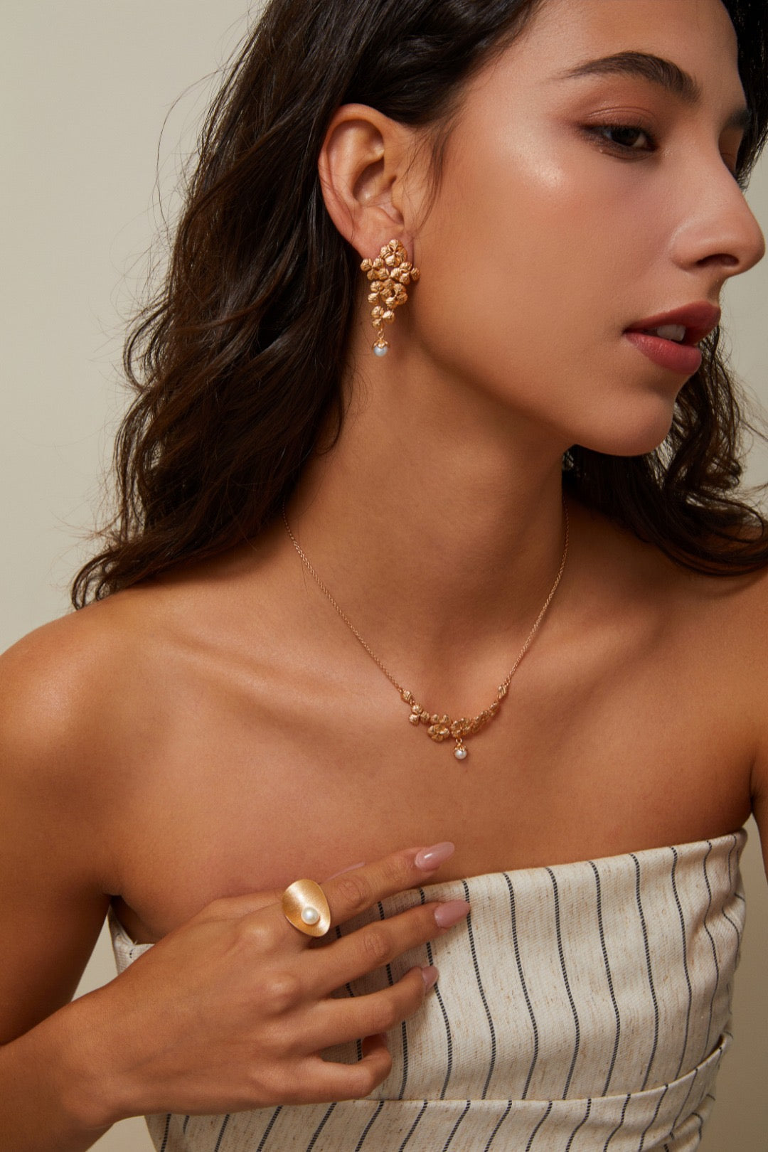Woman wearing gold jewelry including earrings, necklace, and ring against a neutral background