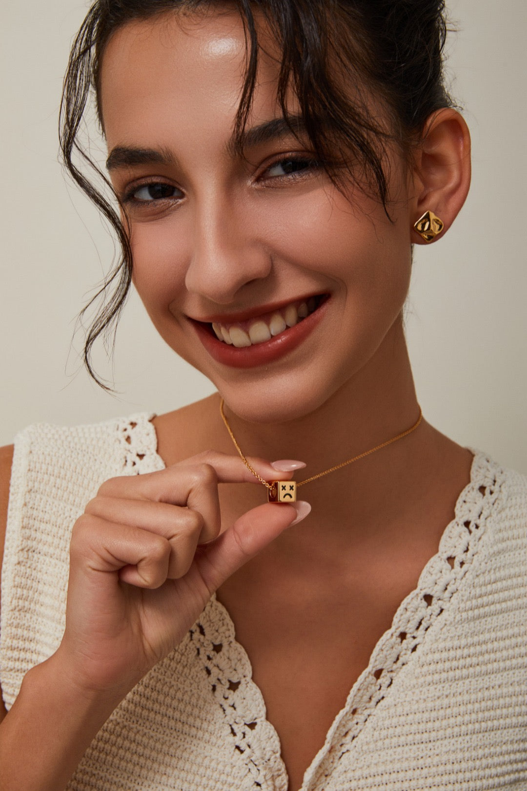 Woman holding a gold necklace with a cube pendant against a neutral background
